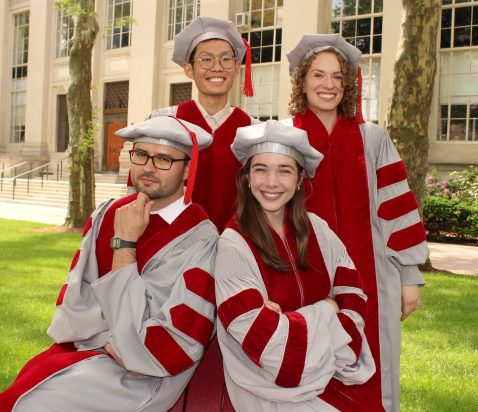 a group of four 2026 PhD graduates in their robes smiling outside the Eastman building on MIT's campus